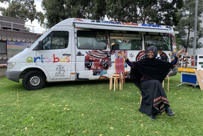 The artsbus mini van parked on green lawn. The van is brightly painted and has seats outside. A lady sits on one on the seats with her arms up, smiling inviting people to participate. 