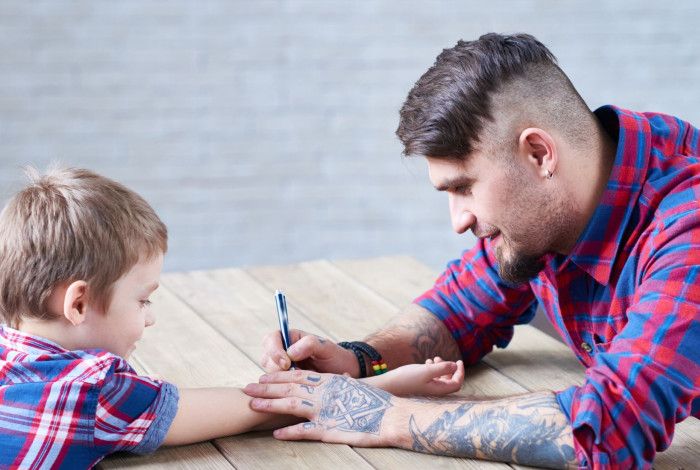 A tattooed adult drawing a temporary tattoo on a child's arm