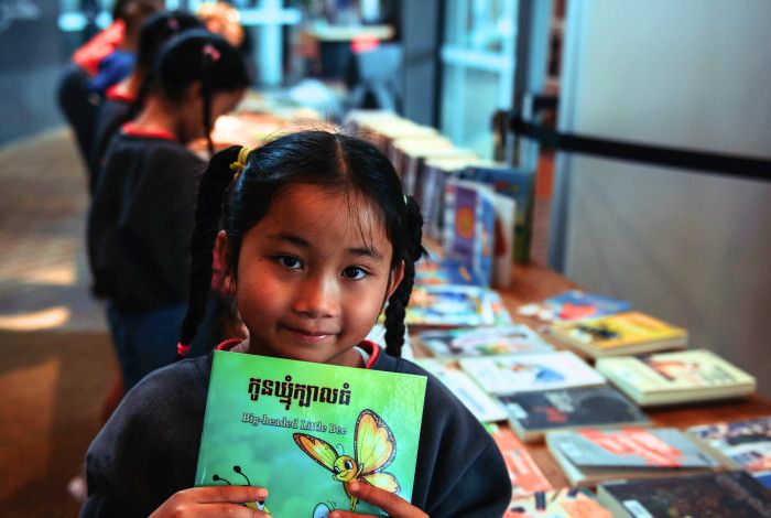 Child holding a library book. There are other children in the background and library books on the table.