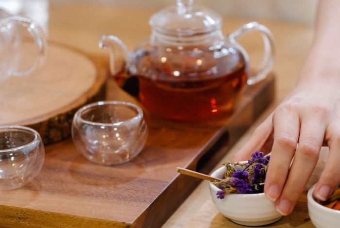 A person selects dried flowers from a small bowl beside a teapot and glass cups during a tea blending session.