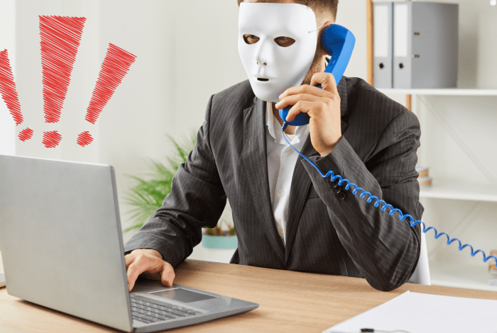 Suspicious looking man in a mask sitting at a laptop on a phonecall.