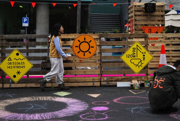 Street scene with wooden barricades decorated with colorful signs and bunting; one person walks past while another kneels drawing chalk art on the pavement.
