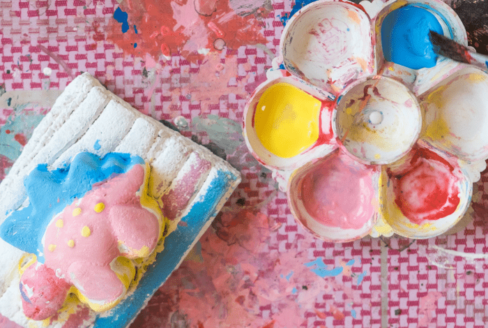 A colourful plaster dinosaur figurine displayed on a pink surface next to a palette with paint samples.