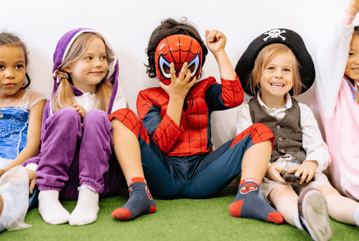 five young children sitting side by side on green carpet wearing costumes. 