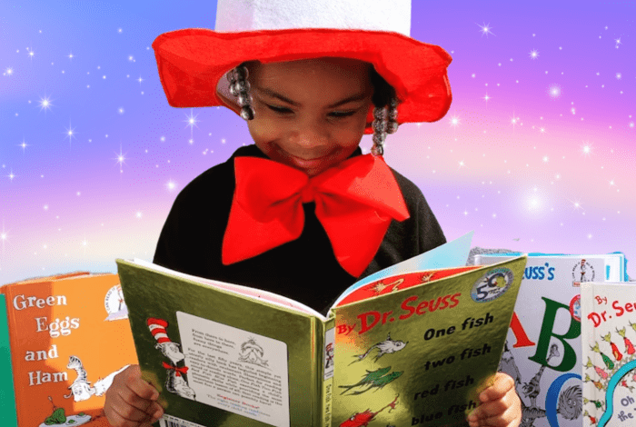 A young girl reading a book, with a smile on her face and dressed in a costume with a large hat and red bow tie.