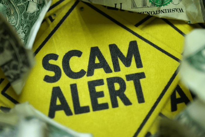 A bright yellow warning sign with words Scam alert surounded by dollar notes.