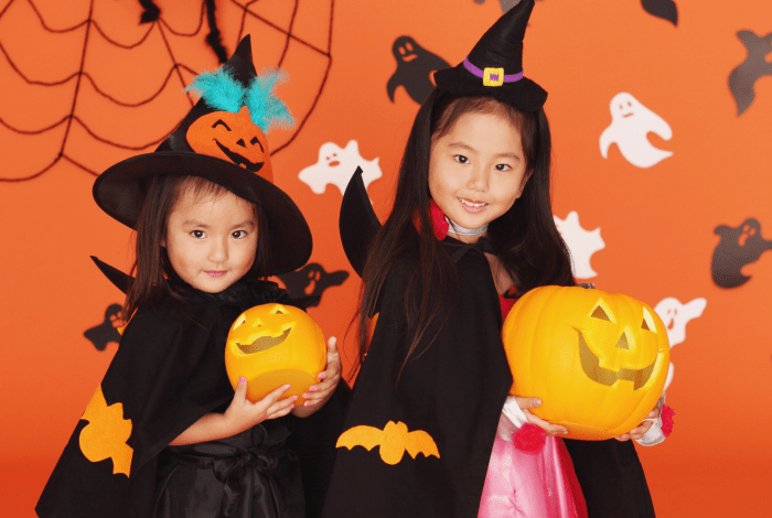 Two children dressed as witches holding pumpkin buckets in front of a Halloween-themed backdrop with ghosts and spider webs.