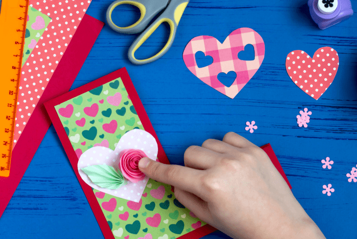 A person holding some colourful craft paper pieces in the shape of love hearts and paper flowers displayed on a blue background together with a range of paper craft paraphernalia.