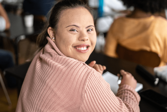 A young girl with special needs, wearing soft pink knitted top and participating in an activity, turned halfway back in her chair and smiling.