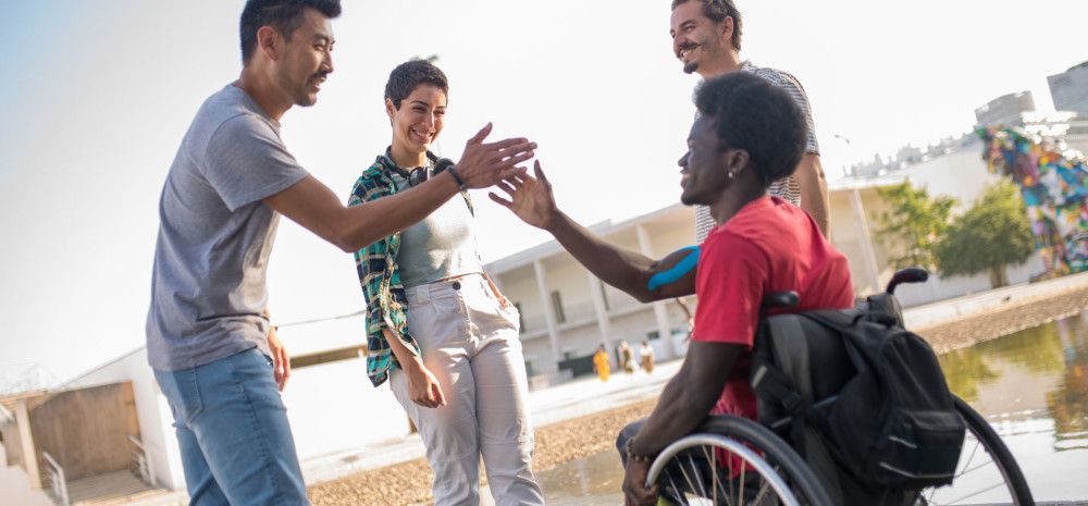 one person is in wheelchair and three other people are standing with smile, one person is giving hi fi to wheelchair person