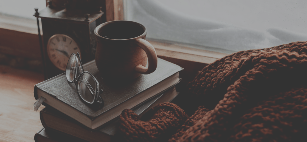 Coffee cup and reading glasses on a pile of books with clock and brown jumper nearby.