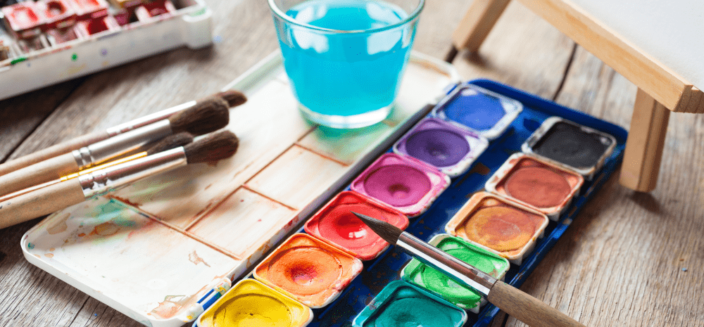 Tray of water colour paints, brushes and glass of water on a table.