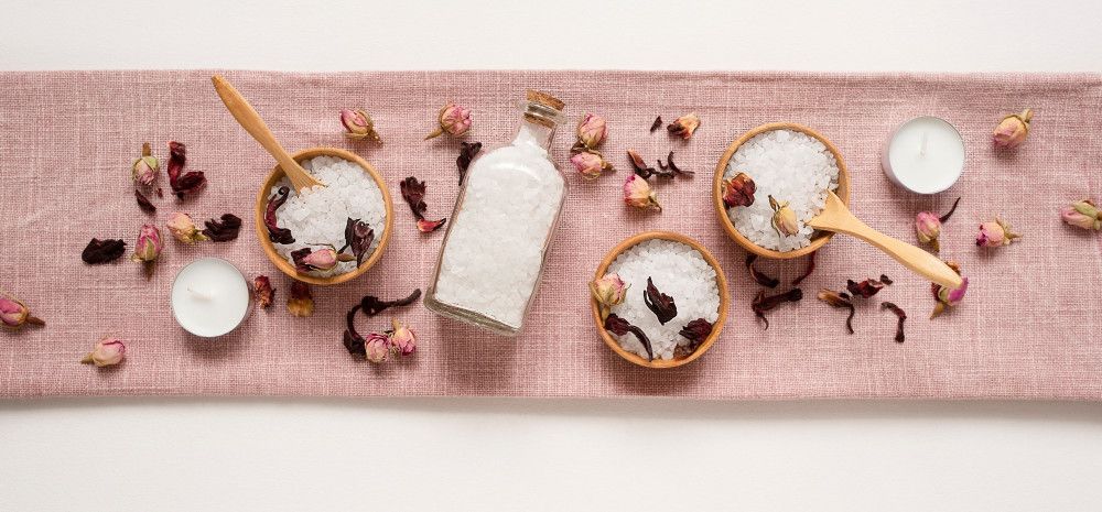 Bath salts in wooden bowls laid out over a pink, petal scattered, table runner.