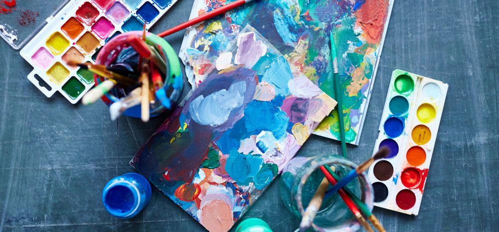 Paints, paintbrushes and artwork on a flat surface.