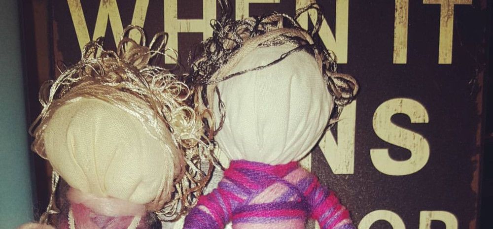 Two handcrafted fabric dolls with yarn hair posed in front of a distressed wooden sign.