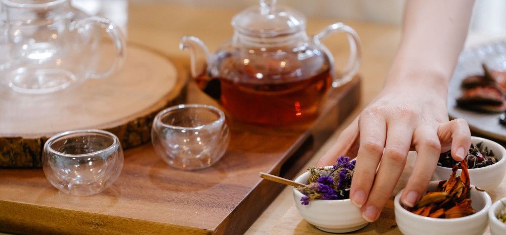A person selects dried flowers from a small bowl beside a teapot and glass cups during a tea blending session.
