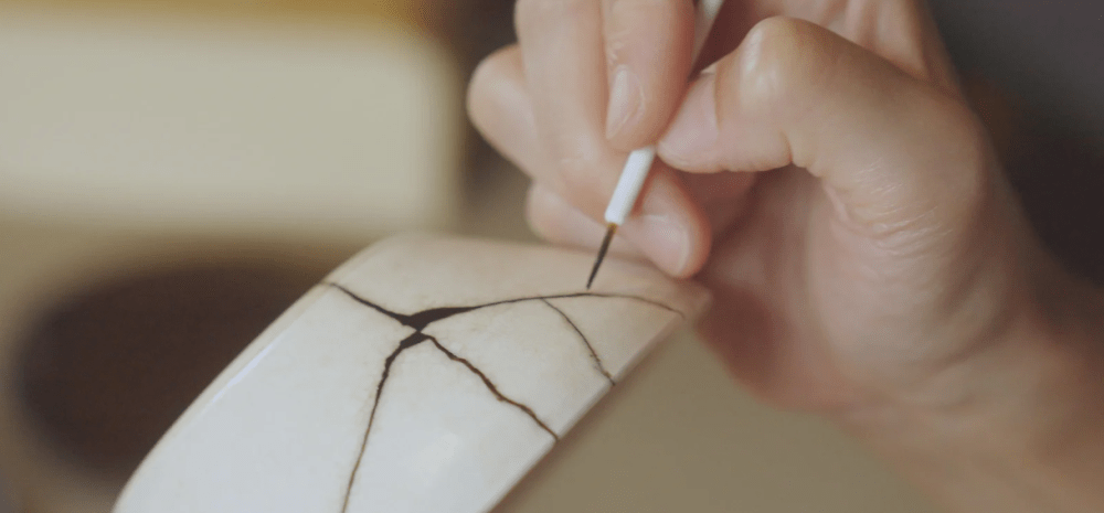 Hand repairing pottery with kintsugi, highlighting cracks with dark lacquer.