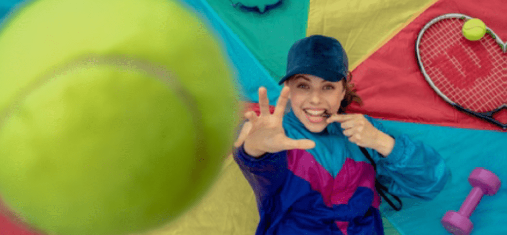 A person in sportswear reaches for a large tennis ball on a colorful mat surrounded by sports equipment
