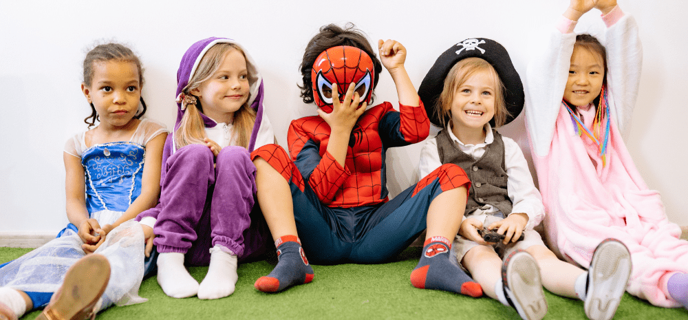 five young children sitting side by side on green carpet wearing costumes. 