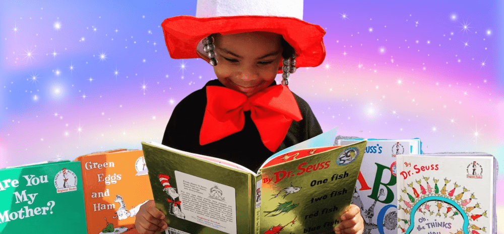 A young girl reading a book, with a smile on her face and dressed in a costume with a large hat and red bow tie.