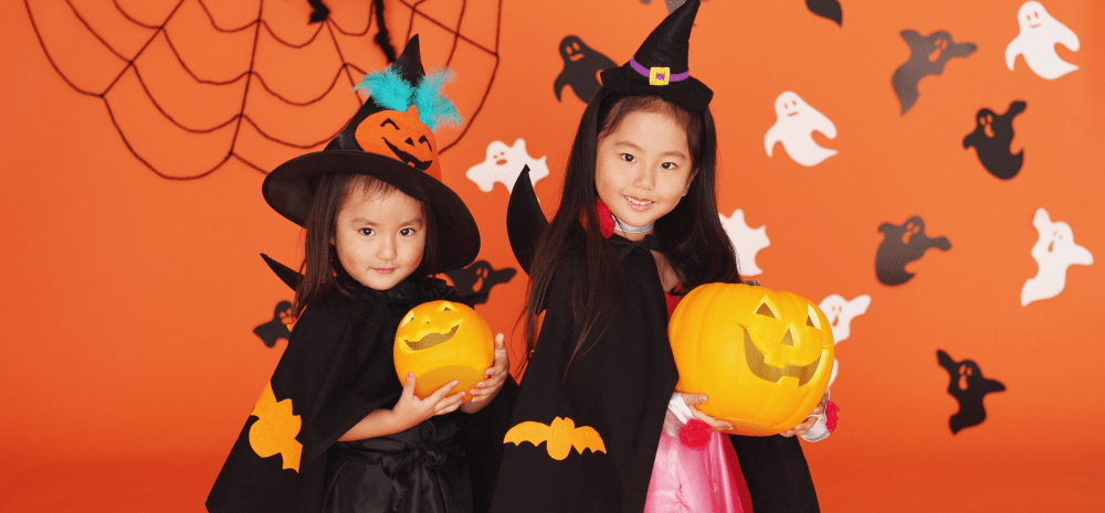 Two children dressed as witches holding pumpkin buckets in front of a Halloween-themed backdrop with ghosts and spider webs.