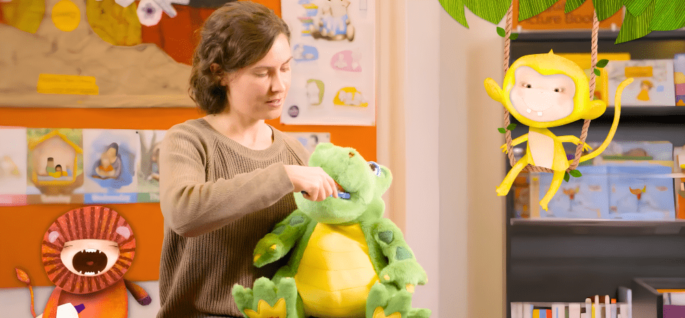 A woman holding a toy dinosaur and brushing his teeth against the background of bookshelves and toys.