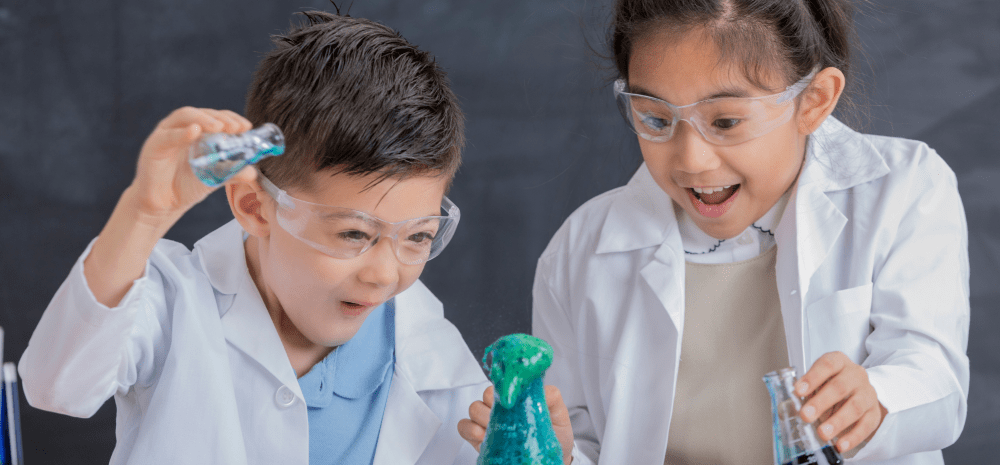 A boy and a girl dressed in white lab coats, wearing safety goggles, holding glass flasks and performing chemical experiments.
