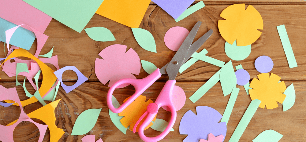 Pink scissors surrounded by colourful paper flowers, leaves and craft cut-outs on a wooden table.