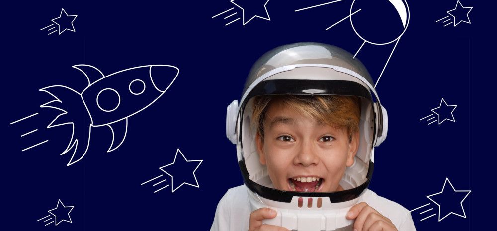 A boy dressed as an astronaut, laughing with excitement, standing in front of a drawing depicting space, rockets and stars.