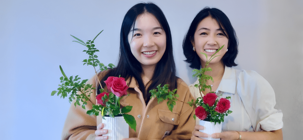 Two Japanese women standing together, facing the camera and holding their ikebana flower arrangements. 