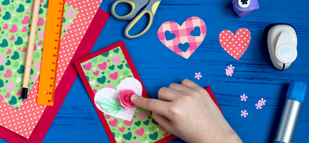 A person holding some colourful craft paper pieces in the shape of love hearts and paper flowers displayed on a blue background together with a range of paper craft paraphernalia.