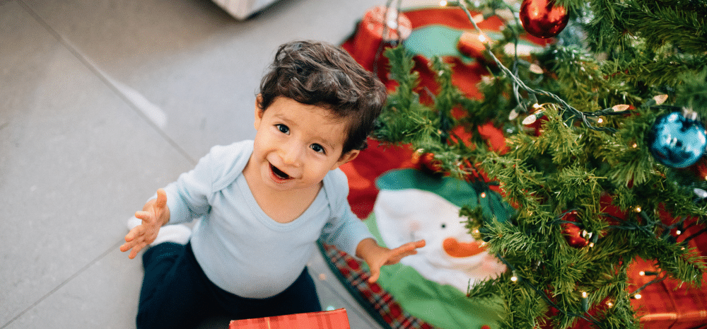 Toddler next to a Christmas Tree, looking up and smiling.