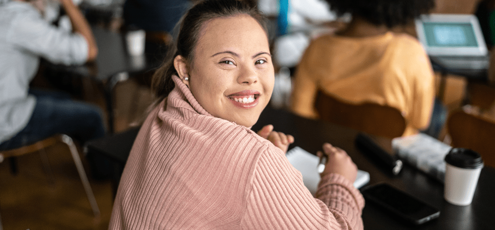 A young girl with special needs, wearing soft pink knitted top and participating in an activity, turned halfway back in her chair and smiling.