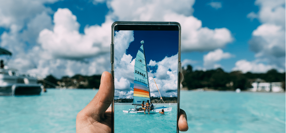 A smartphone frames a colorful sailboat on clear turquoise water beneath a bright sky filled with large white clouds.