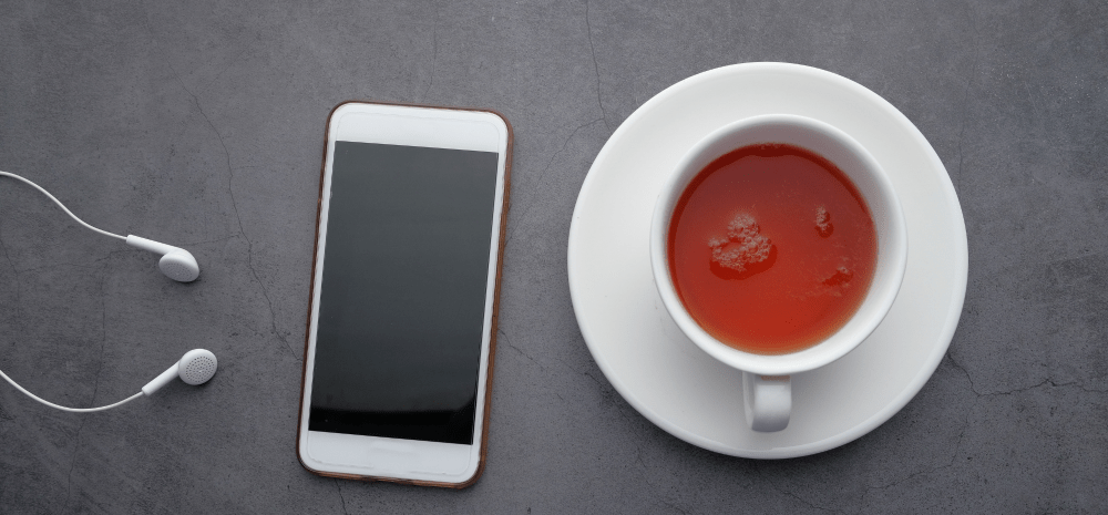 A smartphone, headphones and a cup of tea placed on a gray surface.