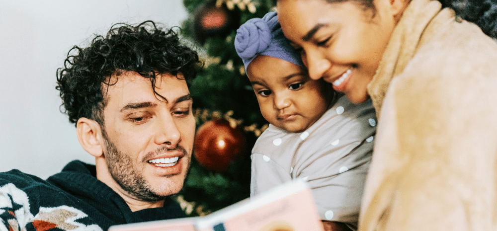 A multicultural family of three standing in front of a Christmas tree and reading a book.