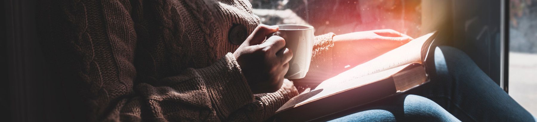 A woman sitting, reading a book and holding a cup in her hand. 