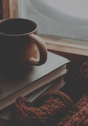 Coffee cup and reading glasses on a pile of books with clock and brown jumper nearby.