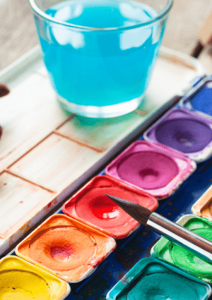 Tray of water colour paints, brushes and glass of water on a table.